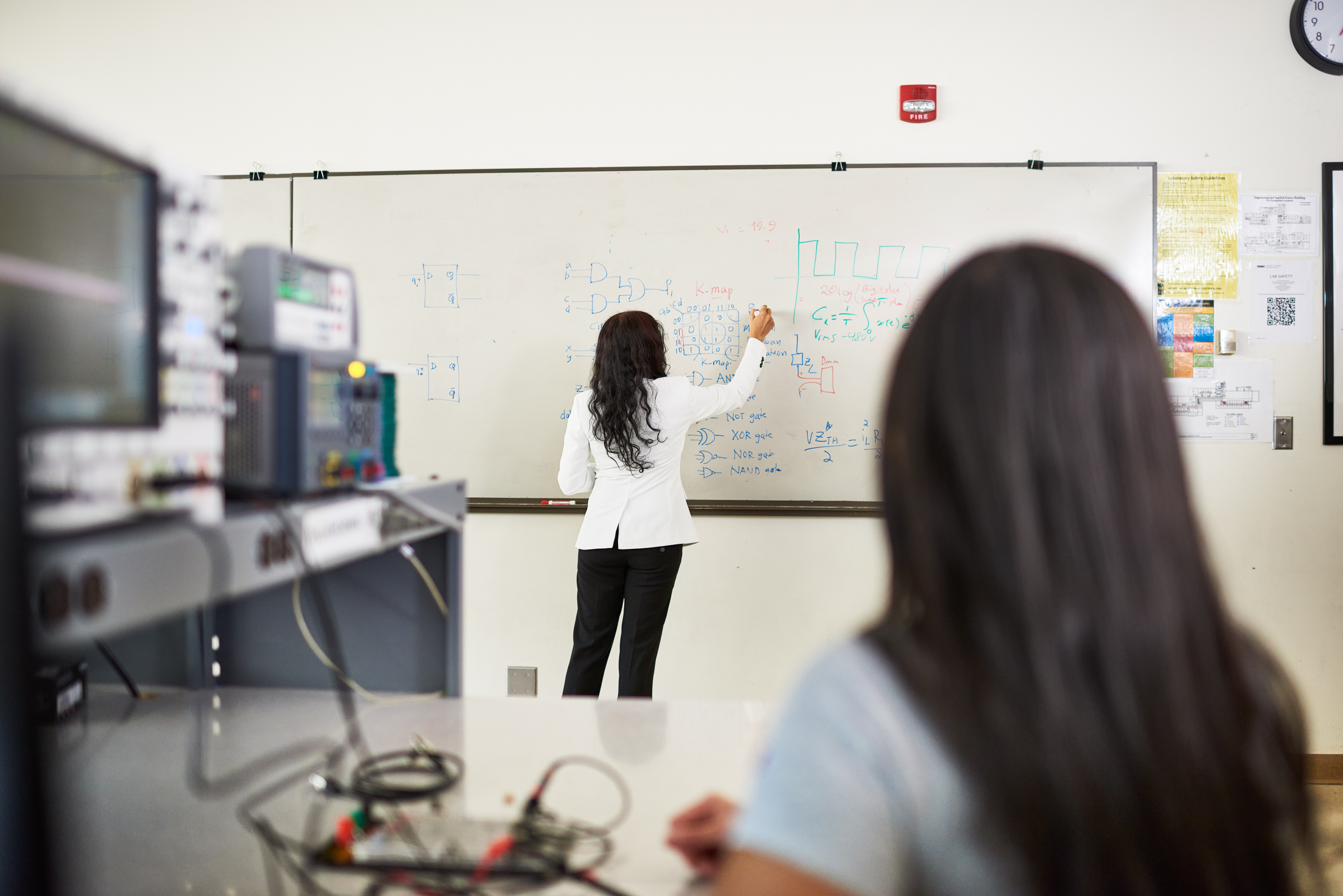 A woman stands at a whiteboard drawing diagrams while a student watches.